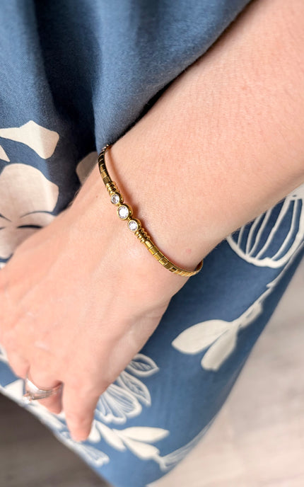 A close-up of a person's wrist wearing a delicate gold bangle bracelet with small white pearl-like beads, set against a blue floral dress.