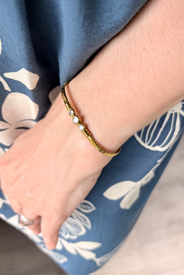 A close-up of a person's wrist wearing a delicate gold bangle bracelet with small white pearl-like beads, set against a blue floral dress.
