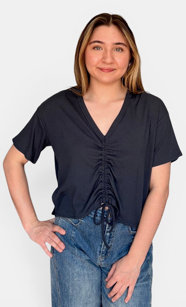 A woman modeling a short-sleeve V-neck top with front ruching and a drawstring tie, paired with blue jeans against a plain light gray background.