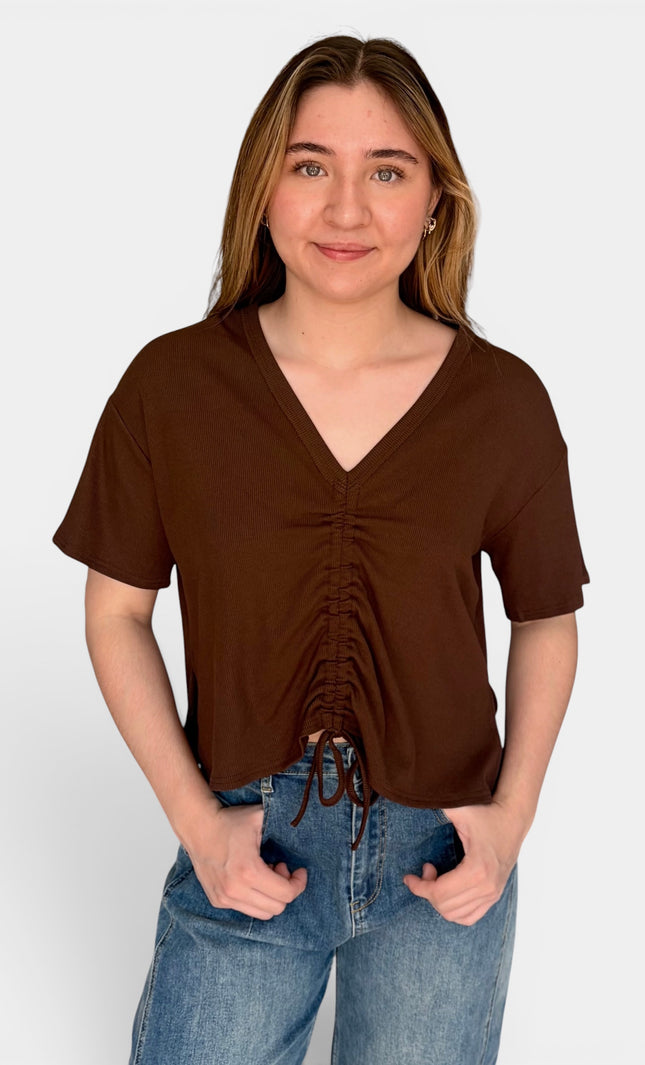 A woman wearing a brown short-sleeve V-neck top with front ruching and drawstring ties, paired with blue jeans, standing against a light gray studio background.