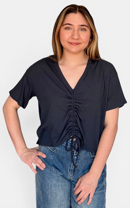 A woman wearing a dark navy blue short-sleeve top with a ruched front and drawstring detail, paired with blue jeans, against a plain light gray background.
