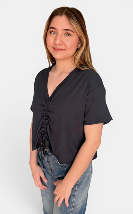 A woman wearing a black short-sleeve top with a ruched front and V-neckline, paired with blue jeans, photographed against a plain light gray background.