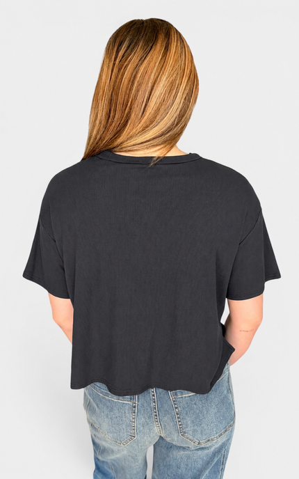 A woman viewed from behind wearing a loose black short-sleeve crop top and light blue jeans against a light gray studio background.