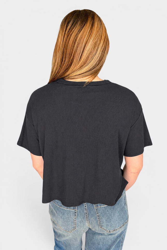 A woman viewed from behind wearing a loose black short-sleeve crop top and light blue jeans against a light gray studio background.