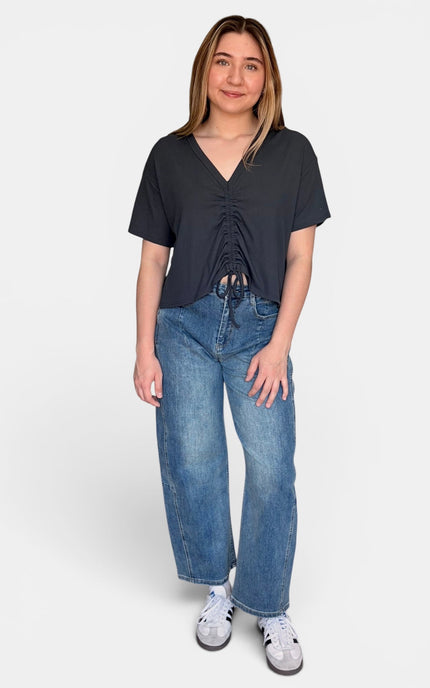 A woman modeling a dark blue short-sleeve top with a ruched front and drawstring detail, paired with blue jeans and white sneakers, against a plain light gray background.