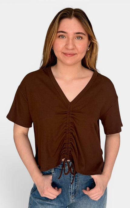 A woman wearing a brown short-sleeve V-neck top with front ruching and drawstring ties, paired with blue jeans, standing against a light gray studio background.