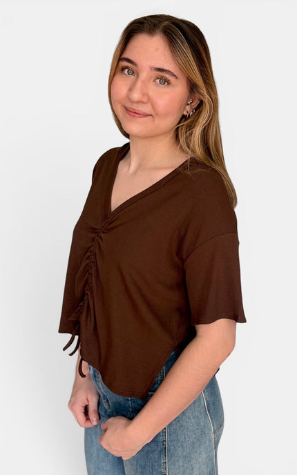 A woman modeling a brown short-sleeve top with a V-neck and side ruching, paired with blue jeans, against a plain light gray background.