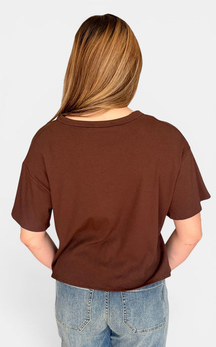 A woman viewed from behind wearing a brown short-sleeve t-shirt and blue jeans against a light gray background.