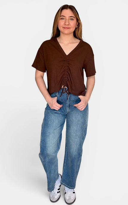 A woman modeling a brown short-sleeve V-neck top with a ruched front and tie detail, paired with blue jeans and white sneakers, against a light gray studio background.