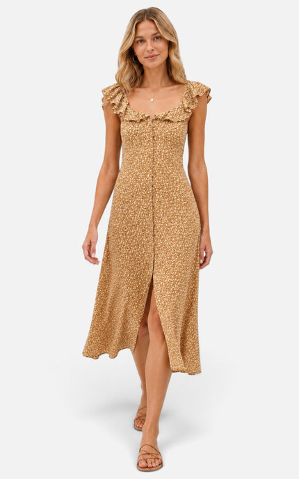 A woman models a sleeveless midi dress with ruffled shoulders and a front slit, against light gray studio background. The dress has a warm tan base with a small repeating floral or ditsy print in a slightly darker golden-brown tone.