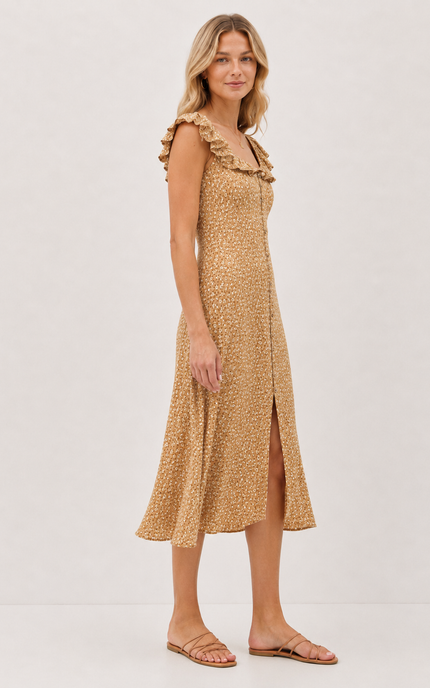 A woman modeling a sleeveless midi dress with ruffled shoulders and a front slit, in a warm tan and brown animal print, against a plain off-white studio background.