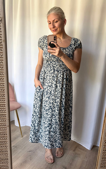 A woman takes a mirror selfie wearing a short-sleeve, scoop-neck maxi dress with a black and white floral pattern. She is standing in front of a white curtain backdrop.