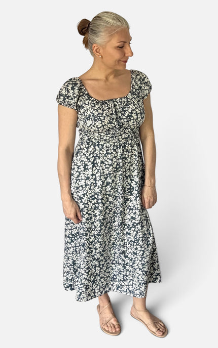 A woman with blonde hair in a bun models a short-sleeved midi dress with a black and white floral pattern, standing against a light gray studio background.