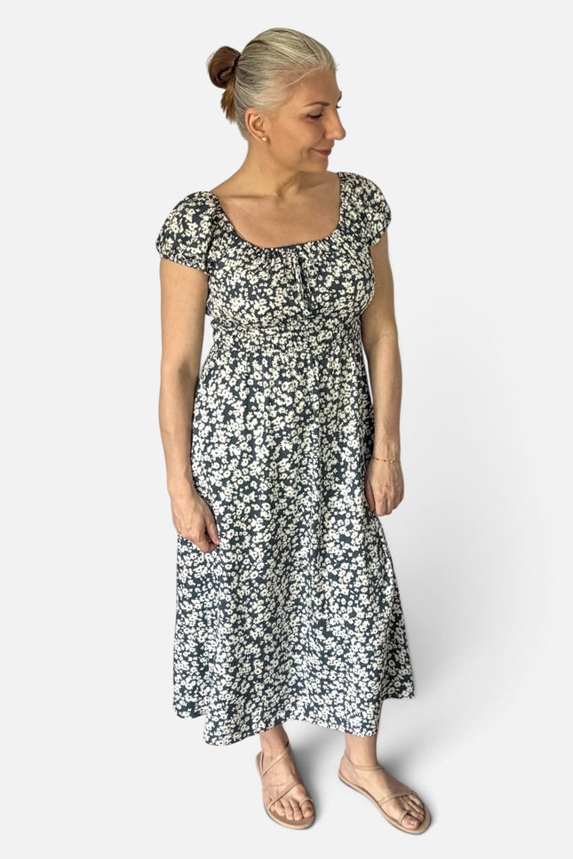 A woman with blonde hair in a bun models a short-sleeved midi dress with a black and white floral pattern, standing against a light gray studio background.