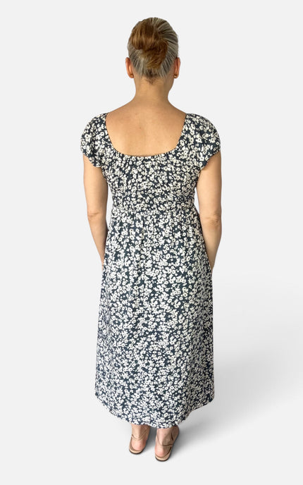 A woman viewed from behind wearing a knee-length floral dress with short cap sleeves and a square neckline, photographed against a light gray studio background.