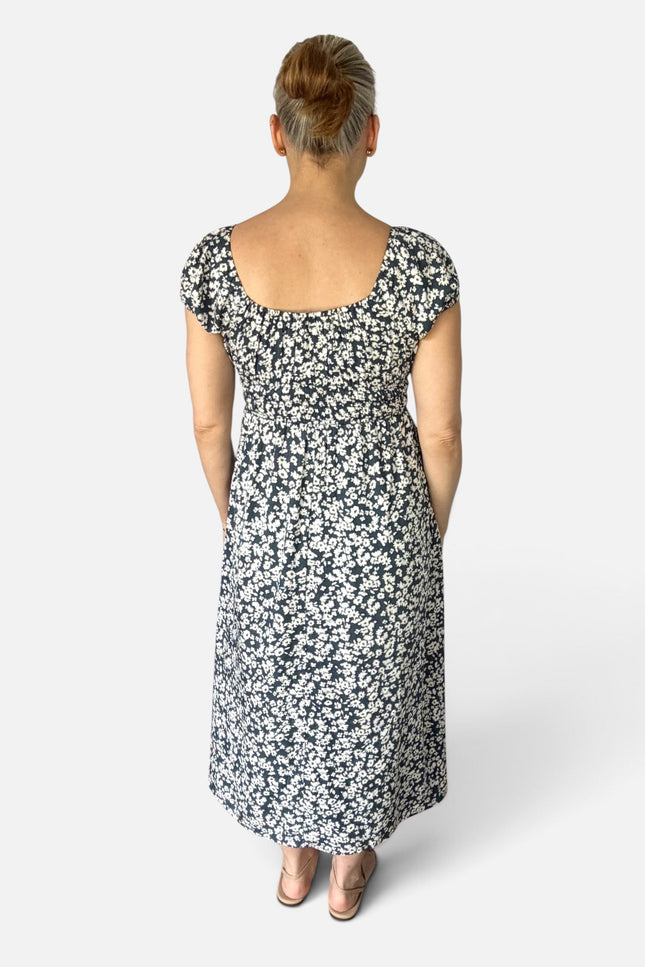 A woman viewed from behind wearing a knee-length floral dress with short cap sleeves and a square neckline, photographed against a light gray studio background.