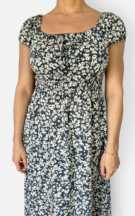 A woman wearing a navy blue midi dress with a dense white floral print, featuring a square neckline with a front tie detail and short puff sleeves, shown against a plain light gray background.