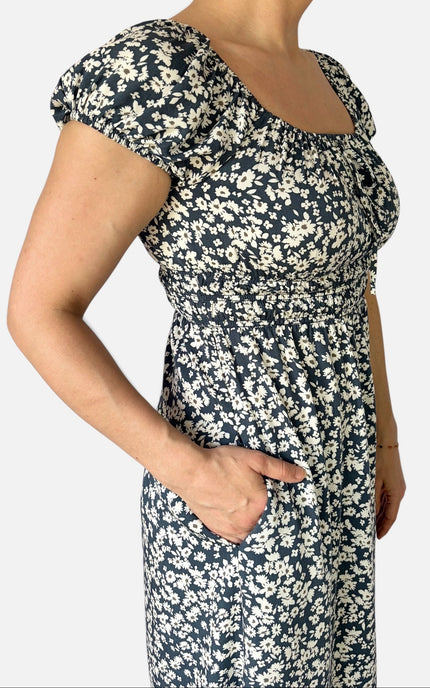 A woman wearing a navy blue midi dress with a white floral print, featuring short puff sleeves, a square neckline, and side pockets, shown against a plain light gray background.