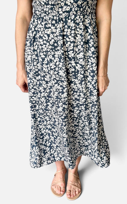 A woman wearing a navy blue midi dress with a dense white floral print, paired with beige flat sandals, shown against a plain light gray background.