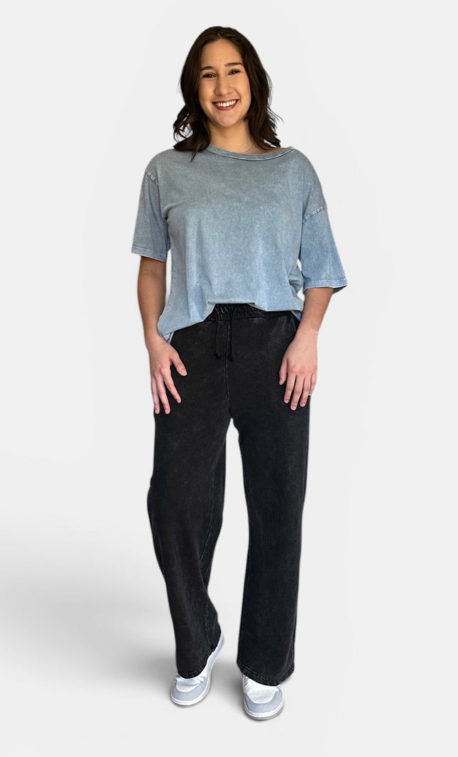 A woman modeling a light blue-gray washed short-sleeve t-shirt and black wide-leg sweatpants against a light gray studio background.