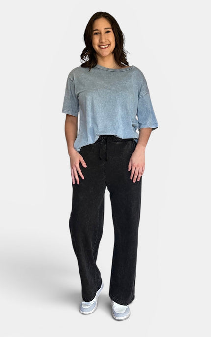 A woman modeling a light blue-gray washed short-sleeve t-shirt and black wide-leg sweatpants against a light gray studio background.
