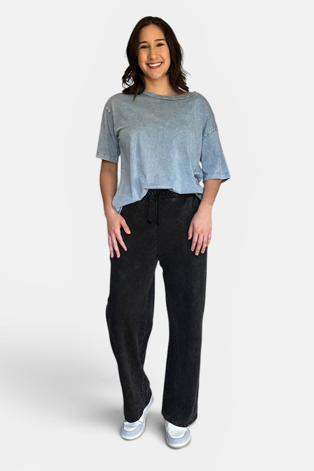 A woman modeling a light blue-gray washed short-sleeve t-shirt and black wide-leg sweatpants against a light gray studio background.