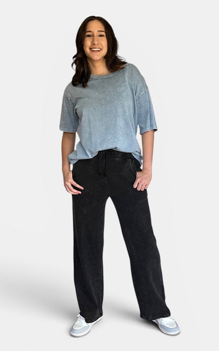 A woman modeling a light blue-gray washed short-sleeve t-shirt and black wide-leg sweatpants against a light gray studio background.