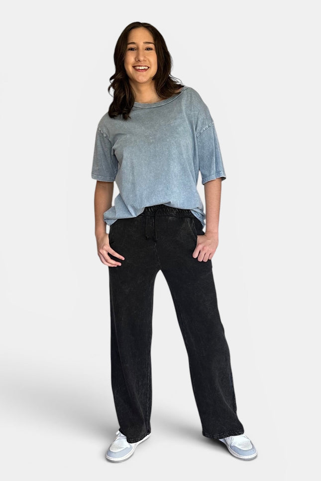 A woman modeling a light blue-gray washed short-sleeve t-shirt and black wide-leg sweatpants against a light gray studio background.