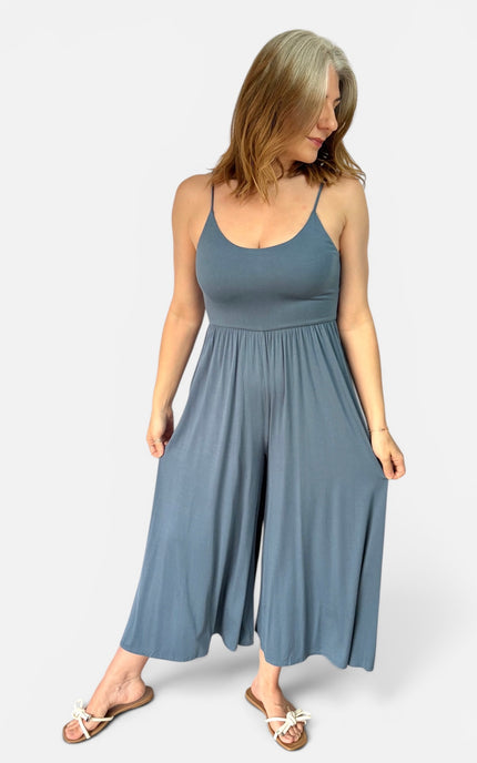 A woman modeling a slate blue sleeveless jumpsuit with wide legs and side pockets, paired with white slide sandals, against a light gray studio background.