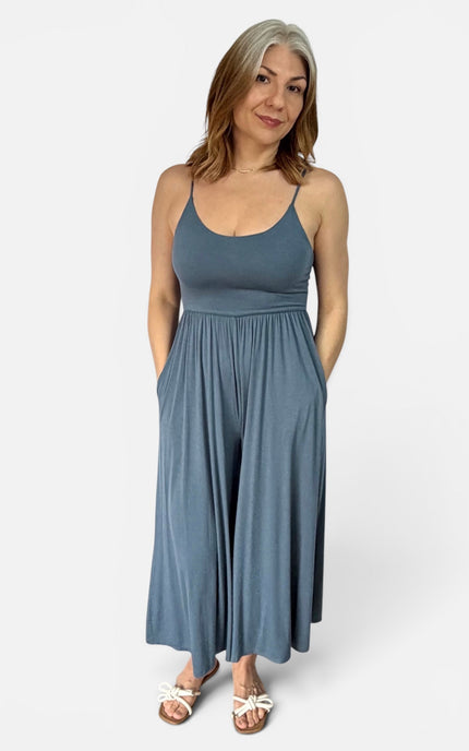 A woman modeling a slate blue maxi dress with spaghetti straps, a scoop neckline, and side pockets, paired with white sandals, against a plain light gray background.