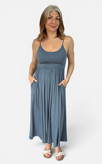 A woman modeling a sleeveless, scoop-neck maxi dress in a dusty slate blue color. The dress features thin spaghetti straps, a gathered waist, and side pockets.