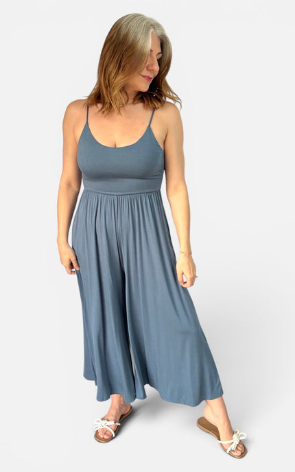 A woman modeling a slate blue sleeveless jumpsuit with a wide-leg, flowing skirt, paired with white slide sandals, against a light gray studio background.