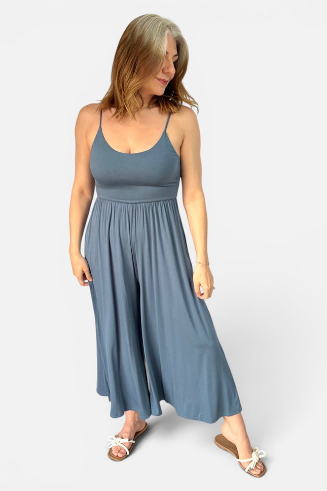 A woman modeling a slate blue sleeveless jumpsuit with a wide-leg, flowing skirt, paired with white slide sandals, against a light gray studio background.
