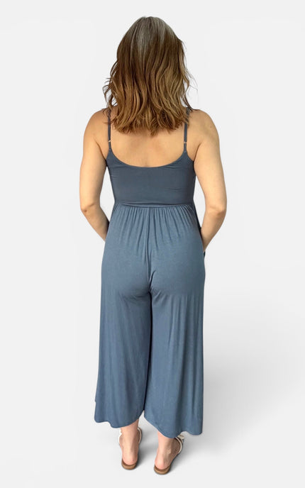 A woman viewed from the back wearing a slate blue sleeveless jumpsuit with thin straps and wide legs, standing against a light gray studio background.
