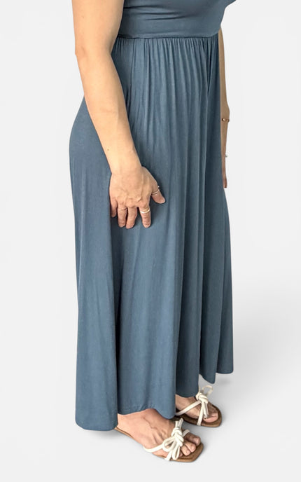 A person wearing a dusty blue maxi skirt with a matching top, paired with white flat sandals, shown from the waist down against a light gray background.