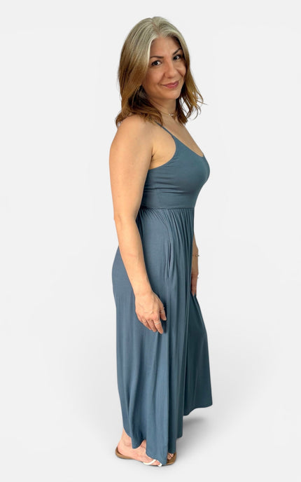A woman modeling a slate blue maxi dress with thin straps and a gathered waist, shown from a side profile against a light gray studio background.