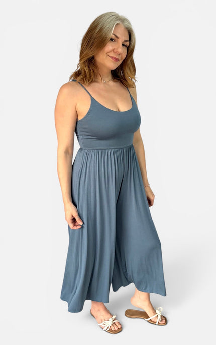 A woman modeling a slate blue sleeveless maxi dress with a scoop neckline and a flowing, gathered skirt, paired with white slide sandals, against a light gray studio background.