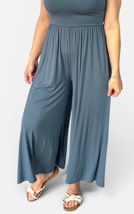 A person wearing a slate blue wide-leg jumpsuit with a gathered waistband, paired with white flat sandals, shown against a light gray studio background.