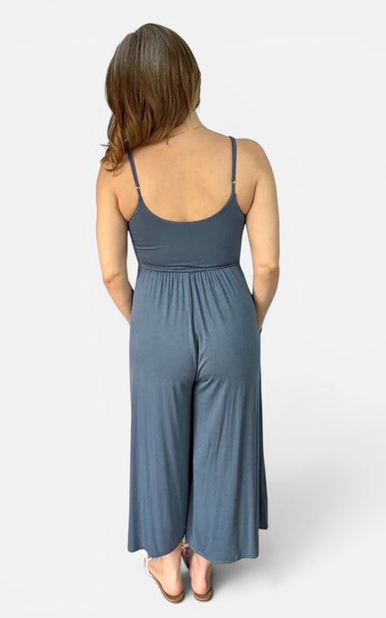 A woman viewed from the back wearing a slate blue sleeveless jumpsuit with thin straps and wide legs, standing against a light gray studio background.