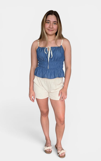 A woman modeling a blue denim-like camisole top with thin straps, a front tie detail, and a gathered peplum hem, paired with cream shorts and sandals, against a light gray studio background.