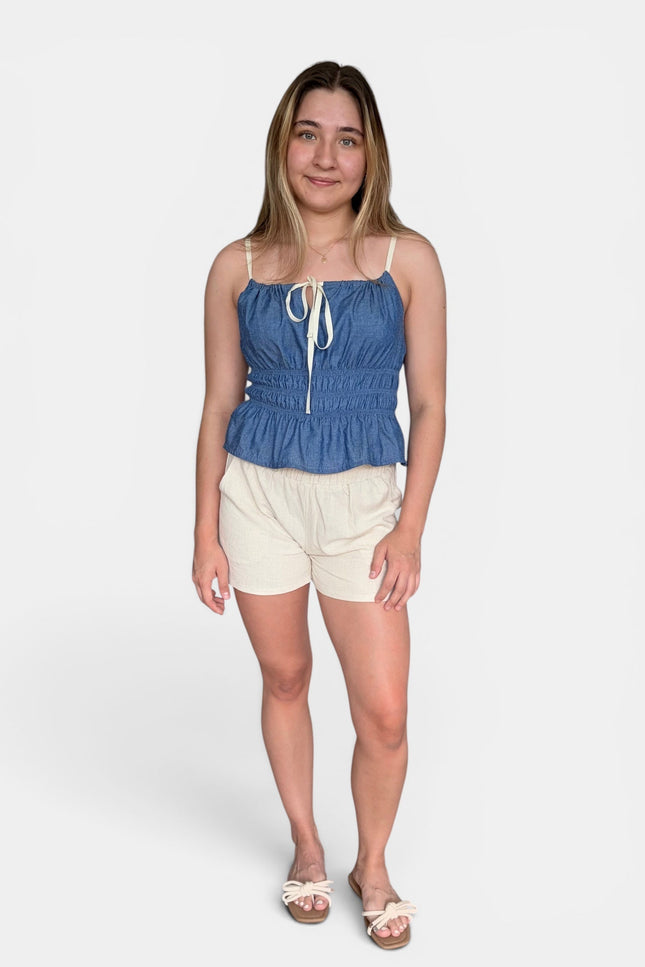 A woman modeling a blue denim-like camisole top with thin straps, a front tie detail, and a gathered peplum hem, paired with cream shorts and sandals, against a light gray studio background.