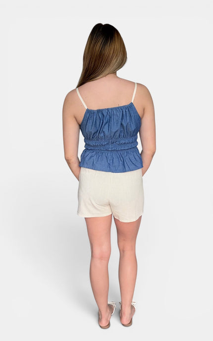A woman shown from the back wearing a blue denim-like camisole top with thin straps and gathered detailing, paired with off-white shorts, against a plain light gray studio background.