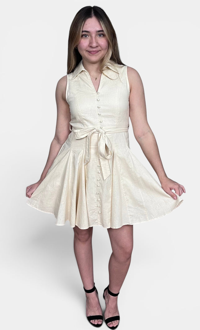 Sleeveless cream-colored mini dress with a button-down front, collar, and matching tie belt at the waist, paired with black high-heeled sandals against a light gray studio background.
