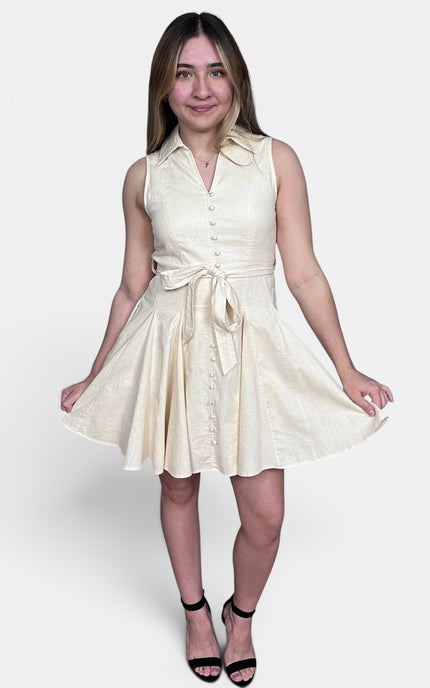 Sleeveless cream-colored mini dress with a button-down front, collar, and matching tie belt at the waist, paired with black high-heeled sandals against a light gray studio background.