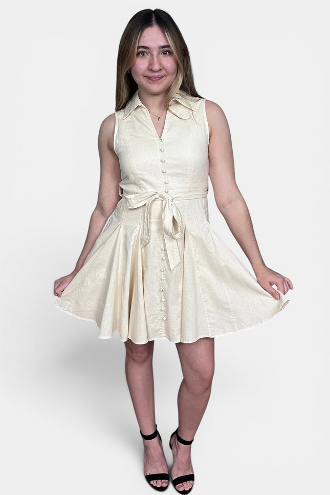 Sleeveless cream-colored mini dress with a button-down front, collar, and matching tie belt at the waist, paired with black high-heeled sandals against a light gray studio background.
