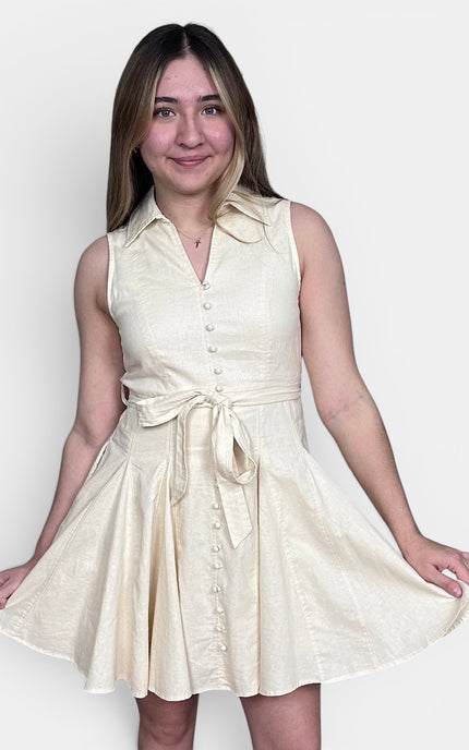 Sleeveless cream-colored mini dress with a tie waist and button front, set against a plain light gray background.