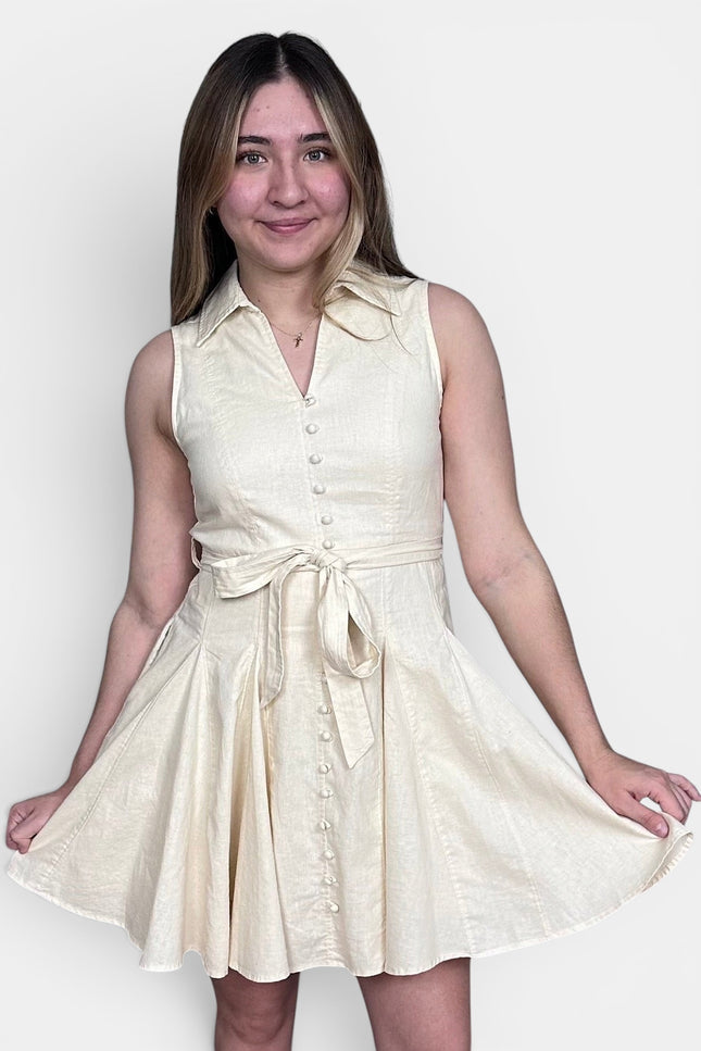 Sleeveless cream-colored mini dress with a tie waist and button front, set against a plain light gray background.