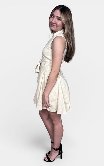 A woman modeling a sleeveless cream-colored mini dress with a tie waist, paired with black strappy high-heeled sandals, set against a plain light gray studio background.