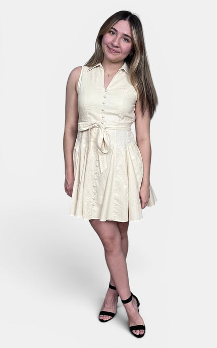 Sleeveless cream-colored mini dress with a button-down front, a matching fabric tie belt at the waist, and a flared skirt, paired with black strappy high-heeled sandals against a light gray studio background.