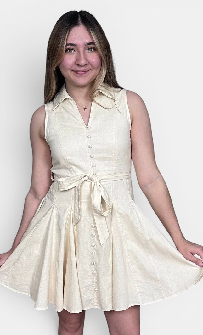 Sleeveless cream-colored mini dress with a tie waist and button front, set against a plain light gray background.